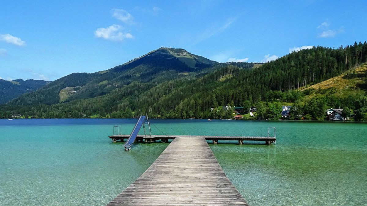 See mit einem Holzsteg, darauf eine Rutsche, türkisblaues Wasser, im Hintergrund einige Häuser und Wald