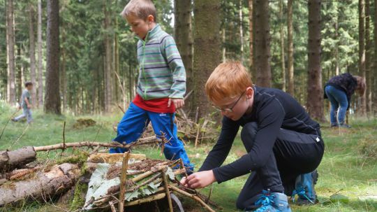 2 Kinder bauen in einem Wald ein Haus aus Rindenstücken