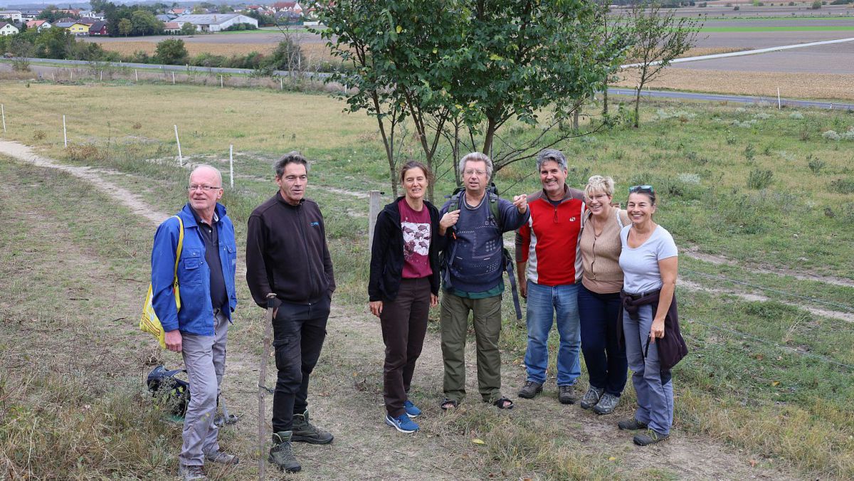 Mehrere Personen stehen auf einem Feldweg vor einem Busch, im Hintergrund Agrarlandschaft.