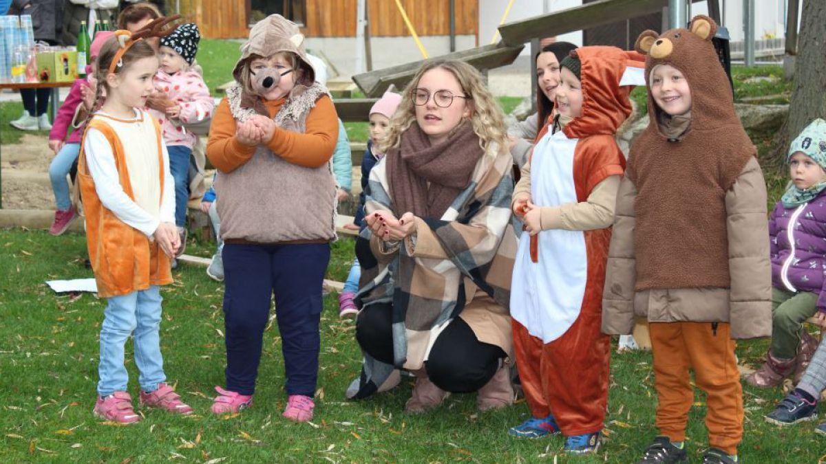 junge Frau kniet mit verkleideten Kindern auf einer Wiese, im Hintergrund sind Spielgeräte und ein Haus zu erkennen