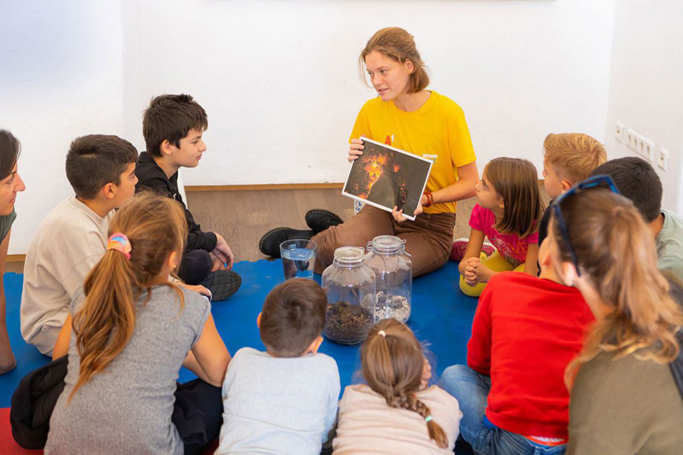 Frau in gelben T-Shirt h&auml;lt eine Schautfale in der Hand, vor ihr sitzen mehrere Kinder in einem Halbkreis auf dem Boden.