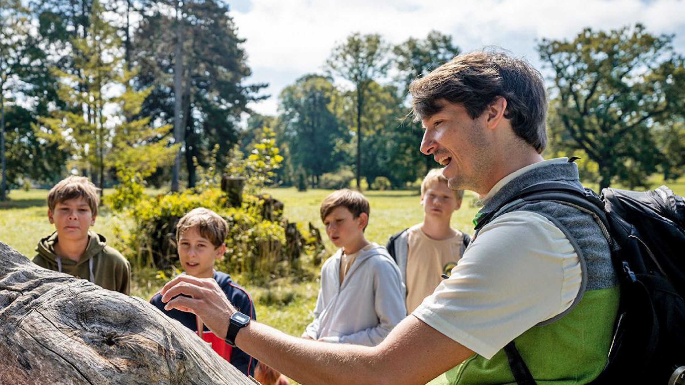 Ein Ranger im Nationalpark Donau-Auen erkl&auml;rt einer Kindergruppe Wissenswertes auf einem Baumstamm. Die Gruppe ist auf einer Wiese, die von Wald begrenzt ist. Es ist strahlender Sonnenschein.