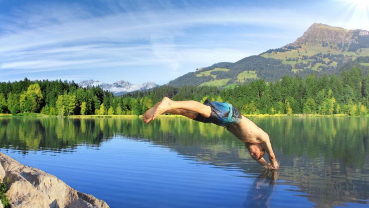 Junger Mann, der einen Kopfsprung in einen See macht, im Hintergrund Wald und Berge, die sich im Wasser spiegeln